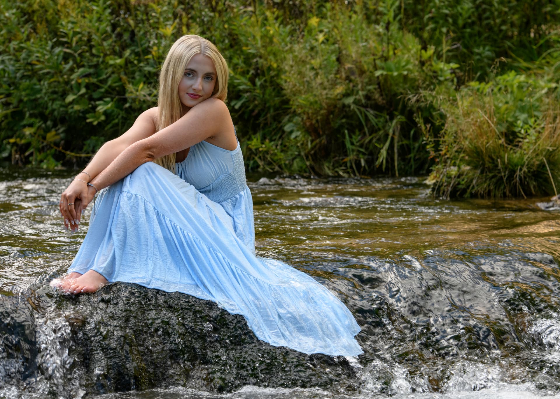 Senior in a flowing light-blue dress seated on a rock in a shallow creek