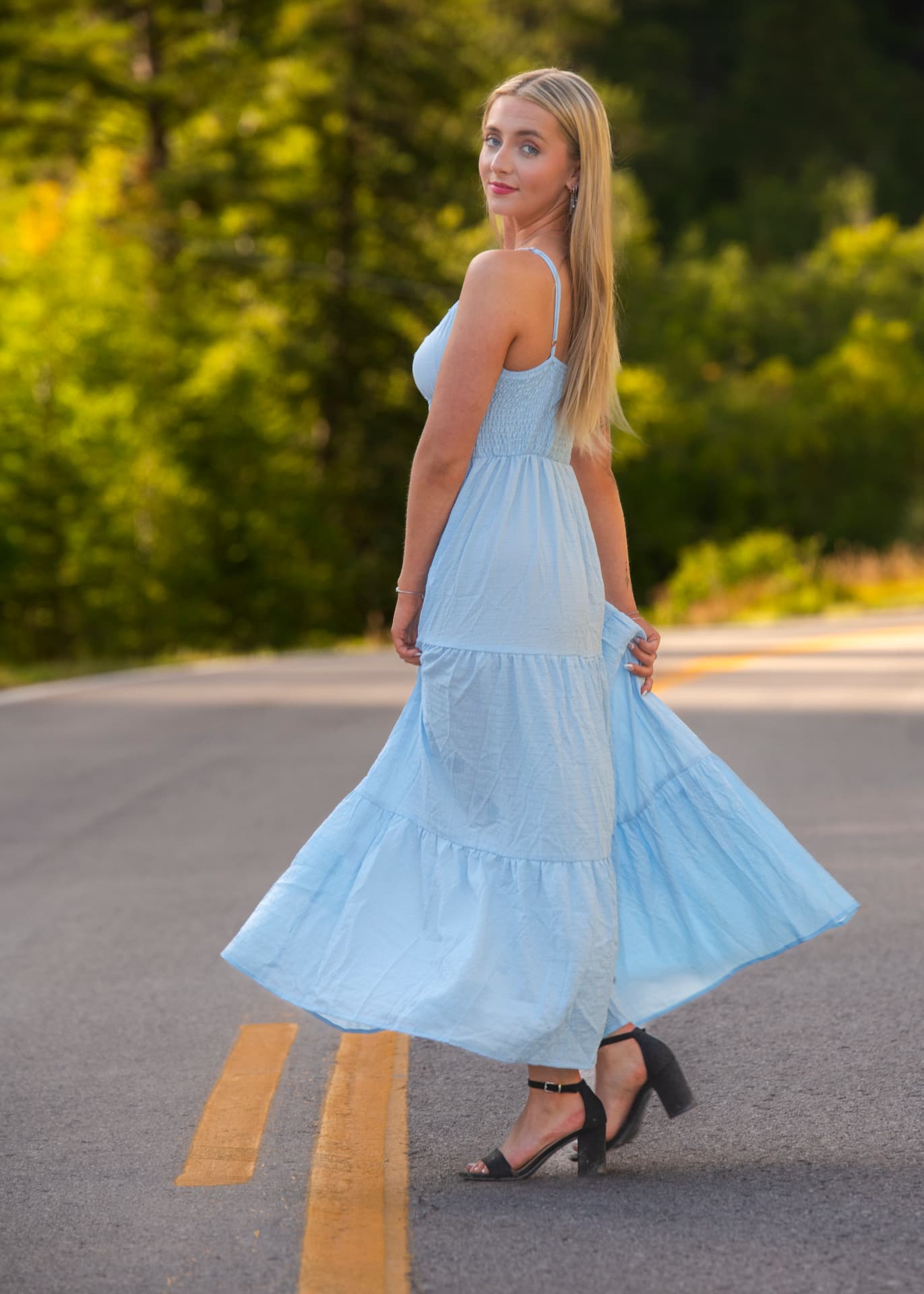 High school senior in a flowing light-blue maxi dress standing on a mountain road lined with sunlit evergreens