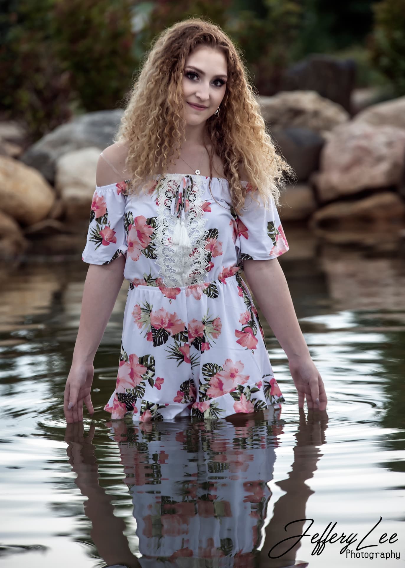 High school senior in a floral off-shoulder dress standing in a calm creek, her reflection in the water