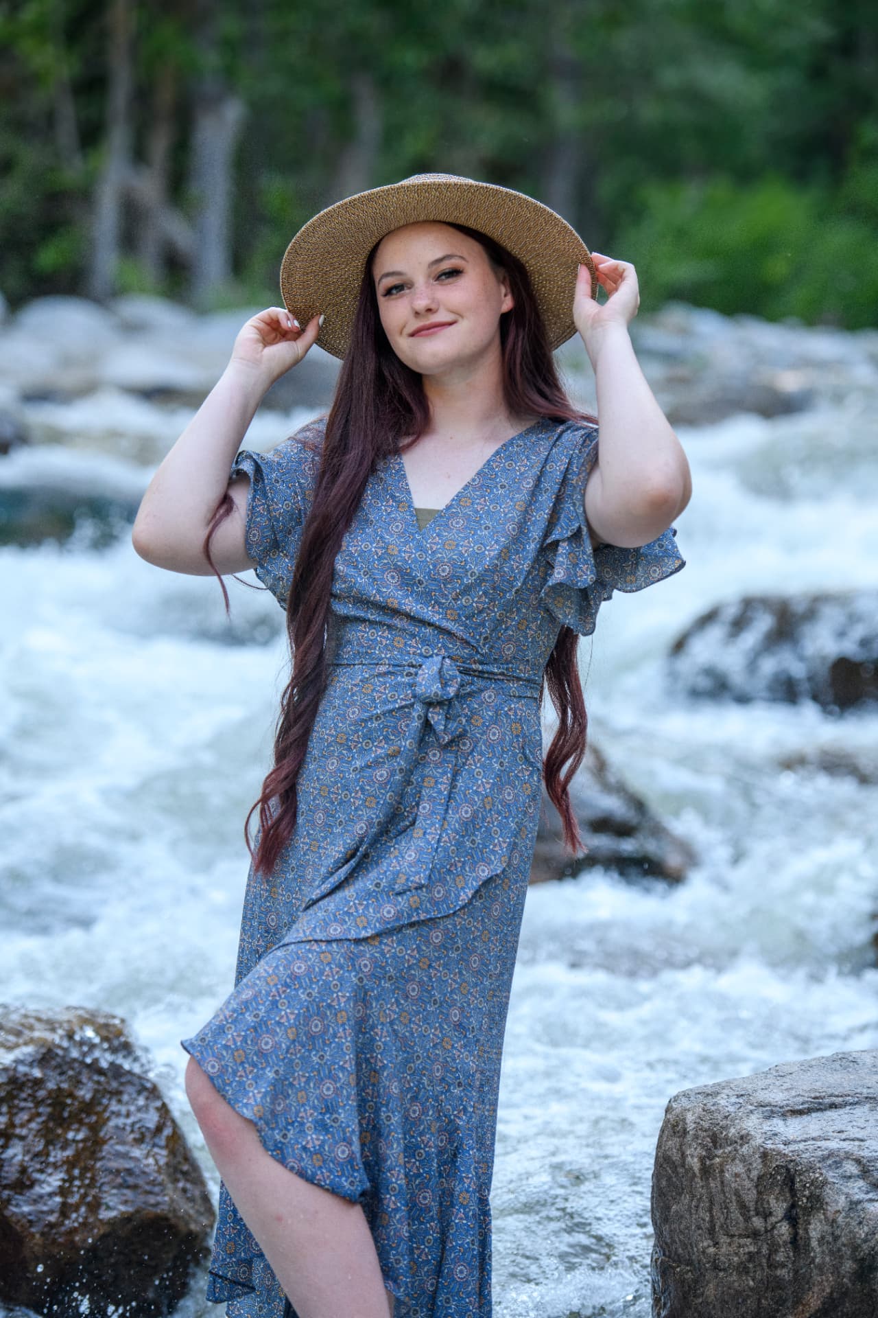 High school senior in a straw hat and blue floral dress standing in a rushing mountain river
